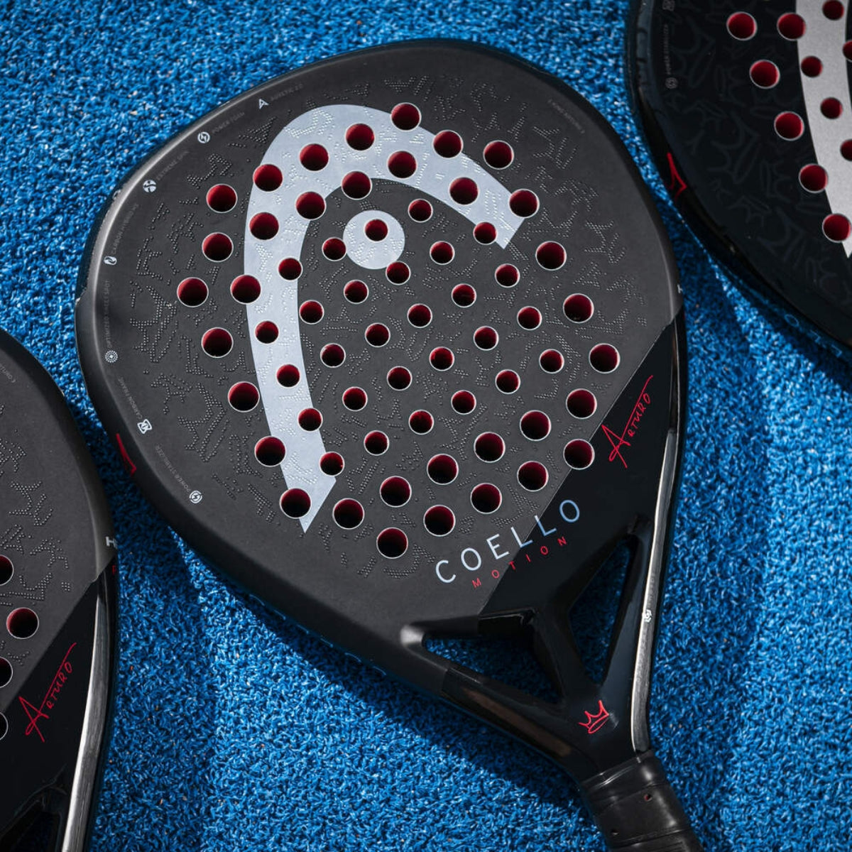 De Head Coello Motion zwarte padel racket met wit Head logo en het woord Coello onderaan, liggend op een blauw padelveld.